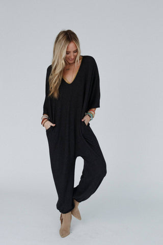 A woman with long blonde hair stands against a light gray background, wearing the Lovely As Ever Harem Jumpsuit in charcoal from Three Bird Nest. The jumpsuit is loose-fitting with a deep V-neckline. She has her hands in the pockets and completes her outfit with tan ankle boots and multiple colorful bracelets. Comfy and boho, she is smiling and looking down.