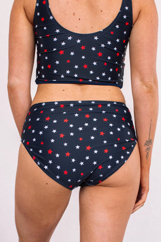 A rear view shows a person wearing the Star Spangled Bottoms from Western Edge Boutique, featuring a two-piece swimsuit adorned with red, white, and blue star patterns. Made from a polyester spandex blend, these stretchy bottoms provide comfort. A small tattoo is visible on their left arm as they pose against a plain white background.