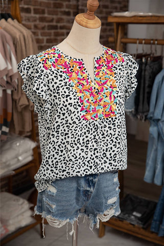 A mannequin dressed in an Aapparella Mac Women Casual Ruffle Embellished V-neck Sleeveless Top, featuring a white and black leopard print adorned with colorful embroidered flowers and ruffled embellishments around the v-neckline. The blouse is paired with distressed denim shorts. The background showcases clothing racks and shelves filled with various garments.