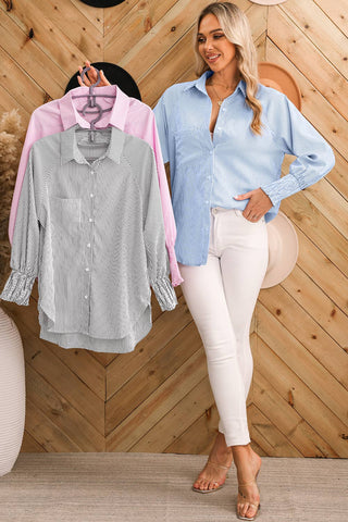 A woman with long blonde hair is posing in front of a wood-paneled wall. She is wearing an Aapparella Taylor Smocked Cuffed Striped Boyfriend Shirt with a turn-down collar, light blue and white stripes, white pants, and beige heeled sandals. In her right hand, she holds pink and grey striped button-up shirts on hangers.
