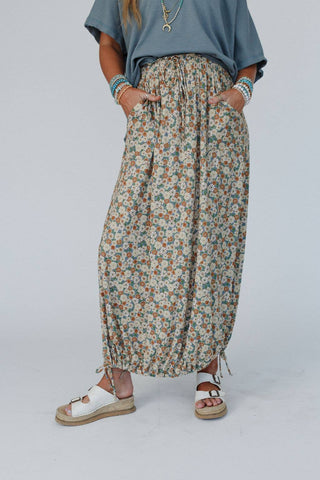 A person wearing a blue top, the Three Bird Nest Solstice Song Cinched Skirt in khaki with pockets, and white platform sandals. The maxi skirt features small multicolored flowers and has drawstrings at the hem. The person accessorizes with several bracelets and has hands in the pockets.