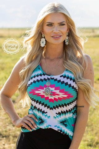 A blonde woman stands outdoors wearing the Stonee Aztec Vibrant Tank by Sterling Kreek, a top adorned with a colorful geometric pattern and crafted from ultra-soft material. She accessorizes with large statement earrings, while her long wavy hair cascades over her shoulders. A tattoo is visible on her left arm as she gazes serenely into the rural landscape behind her.