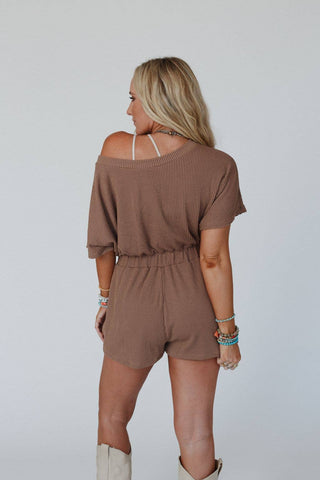 A blonde woman with long hair is seen from the back, wearing a Covey Romper in Camel by Three Bird Nest, which features an elastic waistband and is paired with white boots. She has several colorful bracelets adorning her right wrist. The romper presents a relaxed and casual fit, crafted from ribbed knit fabric. The background is plain and white.