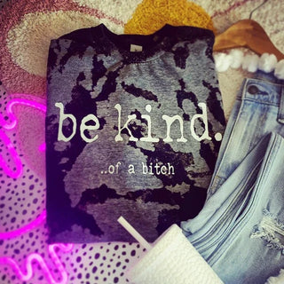 The "BE KIND" shirt by Western Edge Boutique, featuring a camo pattern and the edgy phrase "be kind...of a bitch," boldly stands out against a textured backdrop. Paired with light blue jeans and a white quilted bag, the look is accentuated by a pink neon sign glowing in the background, making for a striking statement.