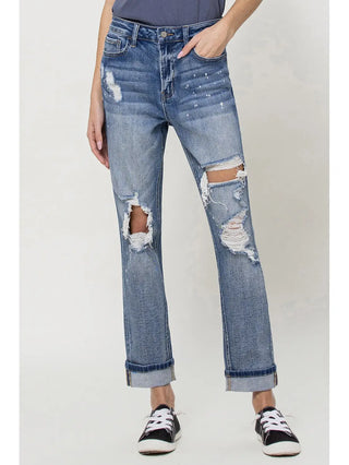 An individual is seen wearing VERVET by Flying Monkey's Abilene Mom Jeans, which showcase distressed detailing and pronounced rips at the knees. The jeans are highlighted with a subtle paint splatter design and are cuffed at the ankles, revealing low-cut black-and-white sneakers. The person adopts a casual pose with one hand in their pocket, paired with a dark-colored top.