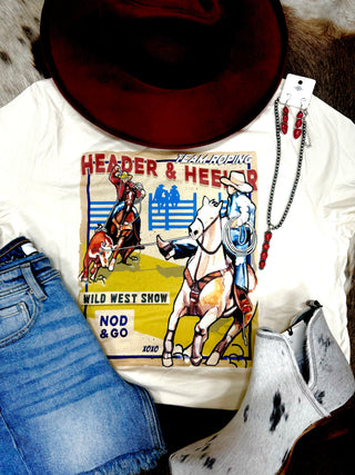 Flat lay of a Western-themed outfit for cowgirl and cowboy enthusiasts, featuring a cream Team Rooing Header & Heeler Graphic Tee from Western Edge Boutique, denim shorts, a red hat, a red beaded necklace, and cow-patterned ankle boots.