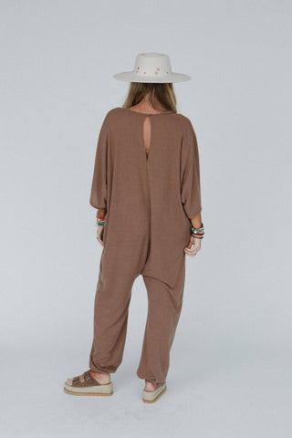 A person with long, straight hair is seen from the back wearing the Lovely As Ever Harem Jumpsuit by Three Bird Nest. The boho, loose-fitting attire features a keyhole opening and is made of comfortable knit fabric. They accessorize with a white wide-brim hat adorned with stars, layered bracelets, and sandals.