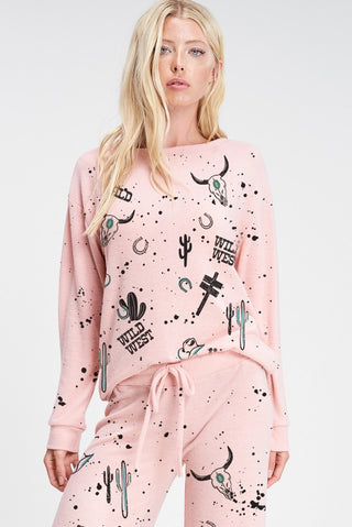 A woman with long blonde hair is wearing the Taylyn Wild West Soft Lounge Set from Phil Love, featuring pastel pink fabric adorned with Wild West-themed prints such as cattle skulls, cacti, horseshoes, and the words "Wild West." The cozy outfit includes a long-sleeve top and drawstring pants. She stands against a white background.