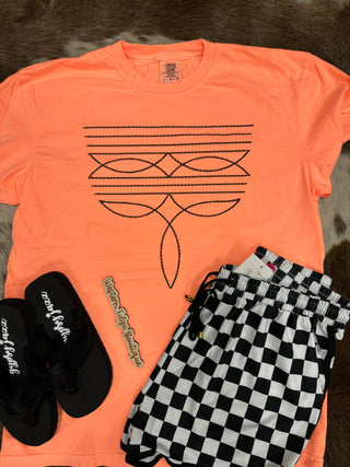 A vibrant orange Tayce Neon Boot Stitch T-shirt from Western Edge Boutique is spread out on a textured surface. Alongside it are stylish black sandals and eye-catching checkered shorts. A small decorative wooden sign enhances the charm of the arrangement.