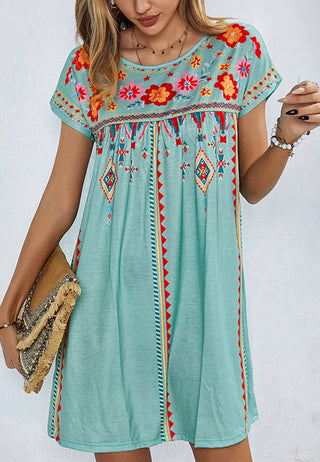 A woman is wearing the Sonnet Multicolor Floral Boho Print Dress by Anna Kaci Wholesale. This short-sleeved turquoise dress features colorful floral and geometric embroidery. She holds a fringed beige clutch bag and accessorizes with bracelets and a necklace. The relaxed fit and vibrant pattern along the neckline and hemline make it perfect for any summer occasion or Sunday dress collection.