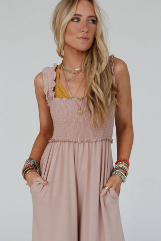 A woman with long, wavy blond hair is standing with her hands in the pockets of a light pink Colt Smocked Ruffle Jumpsuit from Three Bird Nest, featuring sleeveless design and flowy pant legs. She accessorizes with layered necklaces, bracelets, and a visible yellow bra strap.