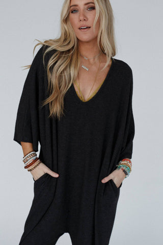 A person with long blonde hair wears the "Lovely As Ever Harem Jumpsuit - Charcoal" by Three Bird Nest and colorful beaded bracelets, exuding a comfy boho style. They stand with hands in their pockets against a plain background, looking directly at the camera.