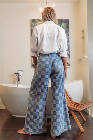 A person stands facing away, dressed in a white shirt and the stylish Wash Two Denim Checkered Jeans by Western Edge Boutique. The wide-leg design with a pleated front adds an elegant touch. They are barefoot on a dark floor, near a white bathtub that is accompanied by a wooden chair, all within a modern, minimalist room.