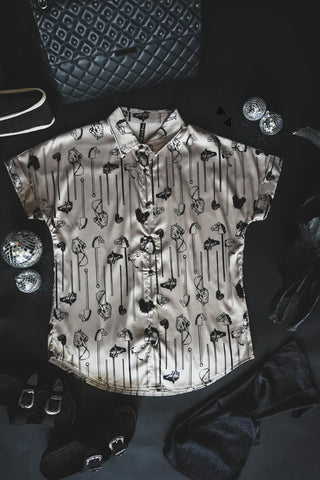 A short-sleeve button-up shirt named "All the Ponies" by 2FLY, featuring a metallic sheen and a pattern of various keys and locks, is laid out on a black surface. Surrounding the retro fit shirt are black velvet boots, a quilted black handbag, a ribbon, and several small disco balls.