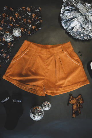 A pair of Buckstitch Queens Saddle mid-rise brown corduroy shorts by 2 Fly, featuring decorative stitching on the pockets, is surrounded by festive items, including silver foil balloons, disco balls, a patterned shirt, black "YEEHAW" socks, and a small cowboy hat decoration.