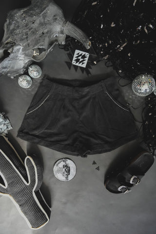 A flat lay image showing the Buckstitch Queen Tux Black mid rise shorts from 2 Fly, decorated with tiny silver studs on the pockets, surrounded by various items. The items include a large skull decoration, shiny ornaments, a black sandal, a knitted cactus, and two disco balls.