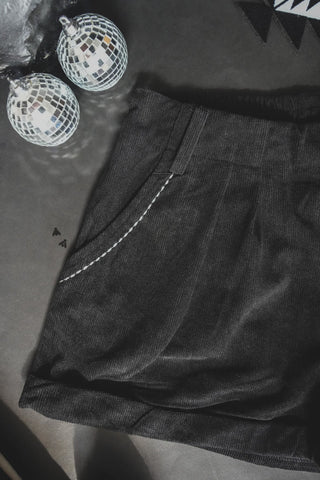 The Buckstitch Queen Tux Black shorts from 2 Fly are laid out on a dark surface. Two small, silver disco ball ornaments are placed nearby, while the background features subtle patterns in black and gray tones.