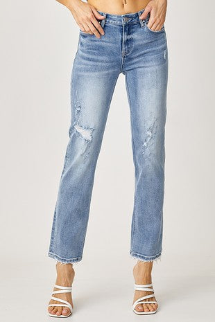 A person models the Risen Baylor Mid Rise Slouch Jeans in light blue, featuring slight distressing and frayed hems. The jeans end just above white, strappy high-heeled sandals. Only the lower torso and legs of the wearer are visible against a plain white background.