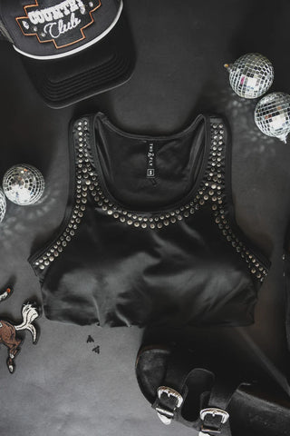 The CABO Cowboy Top by 2 Fly, a black swim top with removable support pads embellished with silver studs around the neckline, is laid out on a black surface. Surrounding items include a black hat featuring the text "Country Club," small silver disco balls, a horse-shaped patch, and a pair of black shoes with silver buckles.