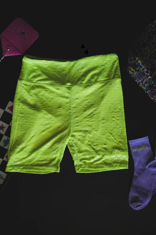 The Chillville Highlighter Biker Shorts from Western Edge Boutique, in a vibrant yellow-green hue, are showcased on a dark surface and made with comfortable stretch material. To the right is a purple sock, while a partially visible patterned bag sits to the left. In the top left corner, there's a pink item adorned with stars.