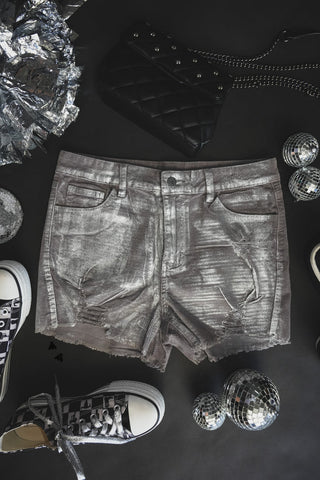 Flat-lay of 2 Fly's COLOR ME CHROME shiny silver denim shorts surrounded by disco balls, black-and-white high-top sneakers, silver wrapping, and a black quilted handbag with a chain strap, all placed on a dark background. The scene exudes a stylish vibe enhanced by the chrome reflections and glittering accessories.