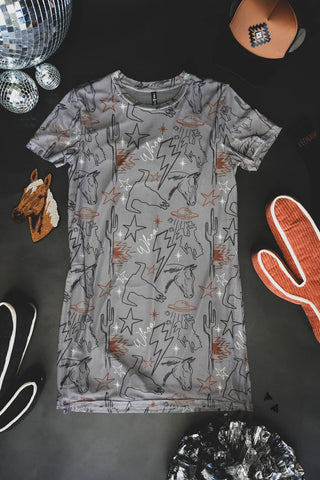 A Cosmic Cowgirl Dress from Western Edge Boutique, showcasing abstract and western-themed designs of horses, cacti, stars, and lightning bolts on a gray T-shirt fabric, is displayed on a dark surface. Surrounding the dress are various items including a disco ball, a hat, orange sneakers, a horse patch, and other accessories. This dress perfectly embodies the spirit of adventurous style.