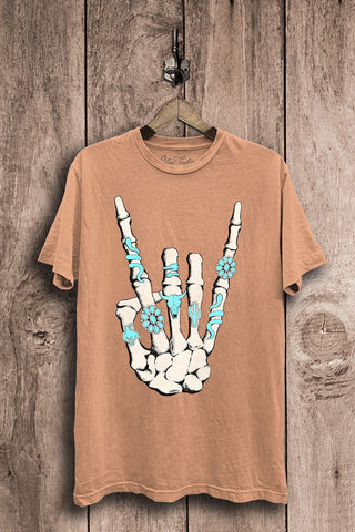 The Dixie Skeleton Rock Hand Graphic Tee from Western Edge Boutique is a tan Mineral Washed Boyfriend style, showcasing a skeletal hand making the 'rock on' gesture with turquoise rings and floral designs. Crafted from 100% cotton, the tee is displayed on a wooden hanger against a rustic wooden background.