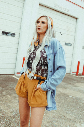 A person with long, light-colored hair stands outdoors. They wear a Buckstitch Queens Saddle by 2 Fly, a graphic t-shirt, a denim jacket, mustard-yellow mid-rise shorts, and a patterned headband. In the background, there's a white garage door flanked by two red posts.
