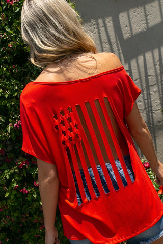 A person with long blonde hair is facing away, wearing the Kooper Red 4th of July Laser Cut-Out Top from Western Edge Boutique, which features a slashed back revealing an American flag pattern and star-shaped cutouts. They are standing in front of a wall partially covered with greenery and flowers.