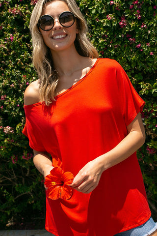 A smiling woman with wavy blonde hair stands in front of a green leafy background with small purple flowers. She wears large round sunglasses and a Kooper Red 4th of July Laser Cut-Out Top from Western Edge Boutique while holding an orange hibiscus flower.