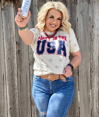A smiling person with wavy blonde hair holds up a can of Michelob Ultra. They are wearing a "Party in the USA" tee from Western Edge Boutique, jeans, and a brown belt, standing in front of a wooden fence.