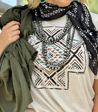 Close-up of a person wearing the TEXAS THREAD Kori Aztec Cross white shirt, featuring an intricate geometric pattern in earthy tones and AZTEC cross-design accents. They have layered silver beaded necklaces, a black bandana with white ovals, turquoise jewelry, and hold an olive rust earthtone jacket over their shoulder.