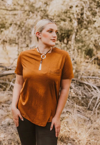 A person with light hair pulled back stands outdoors in a wooded area. They are wearing the HANDY AS A POCKET* BURNT short-sleeve shirt from 2 Fly, made with great fabric and featuring a pocket. They have a western retro accessory around their neck. Their hands are by their sides, and they appear to be looking off to the side.