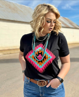 A person with wavy blonde hair stands outdoors wearing the Emerie Black Cheyenne T-shirt from Western Edge Boutique, featuring a colorful Aztec print. They are paired with blue jeans and a brown belt. The individual accessorizes with a layered necklace and wears a watch on their left wrist, looking downwards with their hands in their pockets.