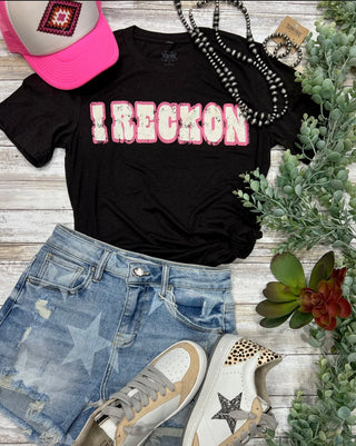 The "I Reckon Tee" by TEXAS THREAD, a classic black T-shirt with "I RECKON" in pink distressed letters, is paired perfectly with light blue denim shorts adorned with a star pattern. Accessories include a pink trucker hat, a black beaded necklace, and white sneakers featuring star designs. Nearby greenery and a succulent complete that perfect country vibe.