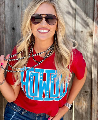 A woman with long blonde hair, wearing sunglasses and the Western Edge Boutique's HOWDY tee featuring bold text, smiles brightly. She holds beaded necklaces against a wooden fence backdrop that perfectly complements her whole cute little number vibe.