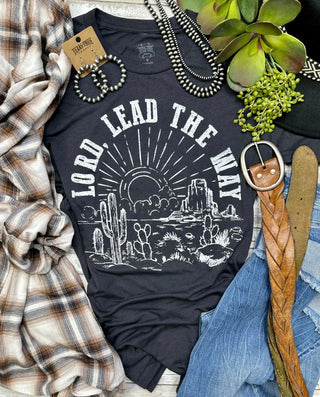 A flat lay image featuring the TEXAS THREAD "Lord Lead The Way" spiritual tee in gray, adorned with the text "LORD, LEAD THE WAY" and a desert scene with cacti. Surrounding the shirt are a plaid shirt, a belt, a pair of earrings, beaded necklaces, a leather strap, and denim pants. Small green plants add to the western vibes.