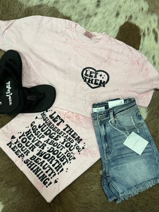 A flat lay showcasing the Western Edge Boutique's LET THEM- Mineral Wash shirt in mineral pink, featuring "Let Them" within a heart, elegantly paired with denim shorts and black sandals on a plush fur rug. The comfort color tee underneath proudly displays the phrase "Let Them Misunderstand You..." for an effortlessly chic appearance.