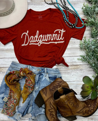 A stylishly knotted brick red unisex Dadgummit Country Wearin Graphic Tee from Western Edge Boutique is paired with distressed denim shorts, a colorful scarf, cowboy boots, a hat, and turquoise necklaces to create an artful ensemble arranged on a rustic wooden surface.