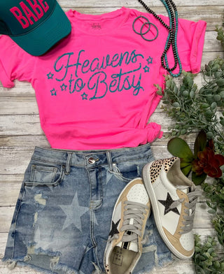 A neon pink t-shirt with the text "Heavens To Betsy Turquoise Ink" by TEXAS THREAD is paired with distressed denim shorts. Accessories include a green cap labeled "Babe," layered beaded necklaces in turquoise hues, and white sneakers featuring leopard print accents and black star details. Succulent plants adorn the background.