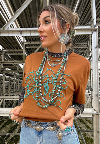 A woman stands in front of a metal structure, wearing a Romee Ranch Road Graphic Tee from 2FLY with a turquoise design. She accessorizes with layered necklaces, turquoise earrings, and a turquoise belt. Her hair is pulled back as she looks to the side, while her light blue jeans are adorned with an embroidered brush type pattern and rings decorate her fingers.