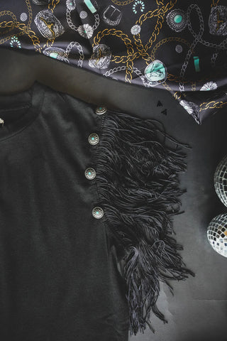 The Get Fringey with it Black Top by 2 Fly, featuring elaborate fringe sleeves and decorative concho details, is laid out on a flat surface. Nearby, a patterned silk scarf adorned with chains and jewels in gold, teal, and white hues, as well as two small disco balls, contribute to a stylish arrangement.