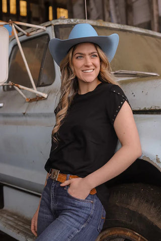 A woman in a CONCHO BOLO BASIC tee from Western Edge Boutique stands smiling beside a rustic vintage truck, her hands resting on her jean pockets, subtly adding to her relaxed demeanor, with the background revealing part of a charming old building.