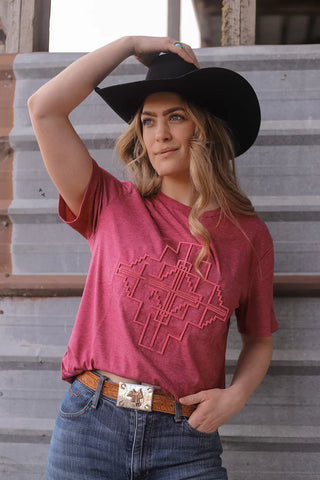 A woman wearing a black cowboy hat, the 2 Fly LAS CRUECES TEE which features an intricate geometric pattern with an embroidered brush technique vibe, a brown belt with a large buckle, and blue jeans. She stands in front of a wooden and metal corrugated wall, posing with one hand adjusting her hat and the other on her hip.