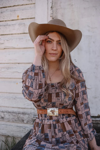 A woman with long blonde hair wearing a wide-brimmed brown hat and the Coastal Cowgirl Dress by 2 Fly stands against a rustic wooden wall. She has her right hand raised, holding the brim of her hat, and she is looking directly at the camera. She also wears a brown belt with a decorative buckle.