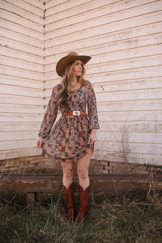 A woman wearing the Coastal Cowgirl Dress by 2 Fly, a brown patchwork design featuring a western check print, stands in front of a weathered white wooden wall. She pairs it with a wide-brimmed hat and cowboy boots. Gazing to the side, one hand rests at her side while the other lightly touches her dress. The ground beneath her is patchy with grass.