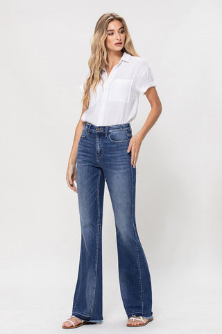 A woman with long, wavy blonde hair wears a white short-sleeve button-up shirt and blue high-waisted Aubrielle High Rise Flare Jeans by VERVET by Flying Monkey, made from comfort stretch denim. She stands against a plain white background, looking slightly to her left, with one hand on her hip and the other hanging by her side. She is also wearing sandals.