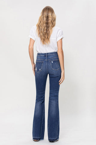 A person with long, wavy hair is wearing a white short-sleeve shirt tucked into high-waisted, dark blue Aubrielle High Rise Flare Jeans from VERVET by Flying Monkey, made of comfort stretch denim. They are standing with their back to the camera against a plain white background.