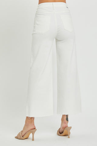 Back view of a person wearing Risen's Winslow Curvy High Rise Tummy Control Crop Wide Leg Pants. The person is standing with feet apart, showcasing the chic design of these white, high-waisted, frayed-hem pants. They're paired with beige open-toe heels against a plain white background.