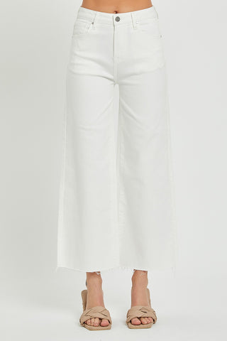 A person is wearing Risen's Winslow Curvy High Rise Tummy Control Crop Wide Leg Pants in white, featuring a frayed hem. These stylish jeans provide excellent tummy control and comfort. They are also wearing beige cross-strap slide sandals. The photo is taken from the waist down against a plain white background.